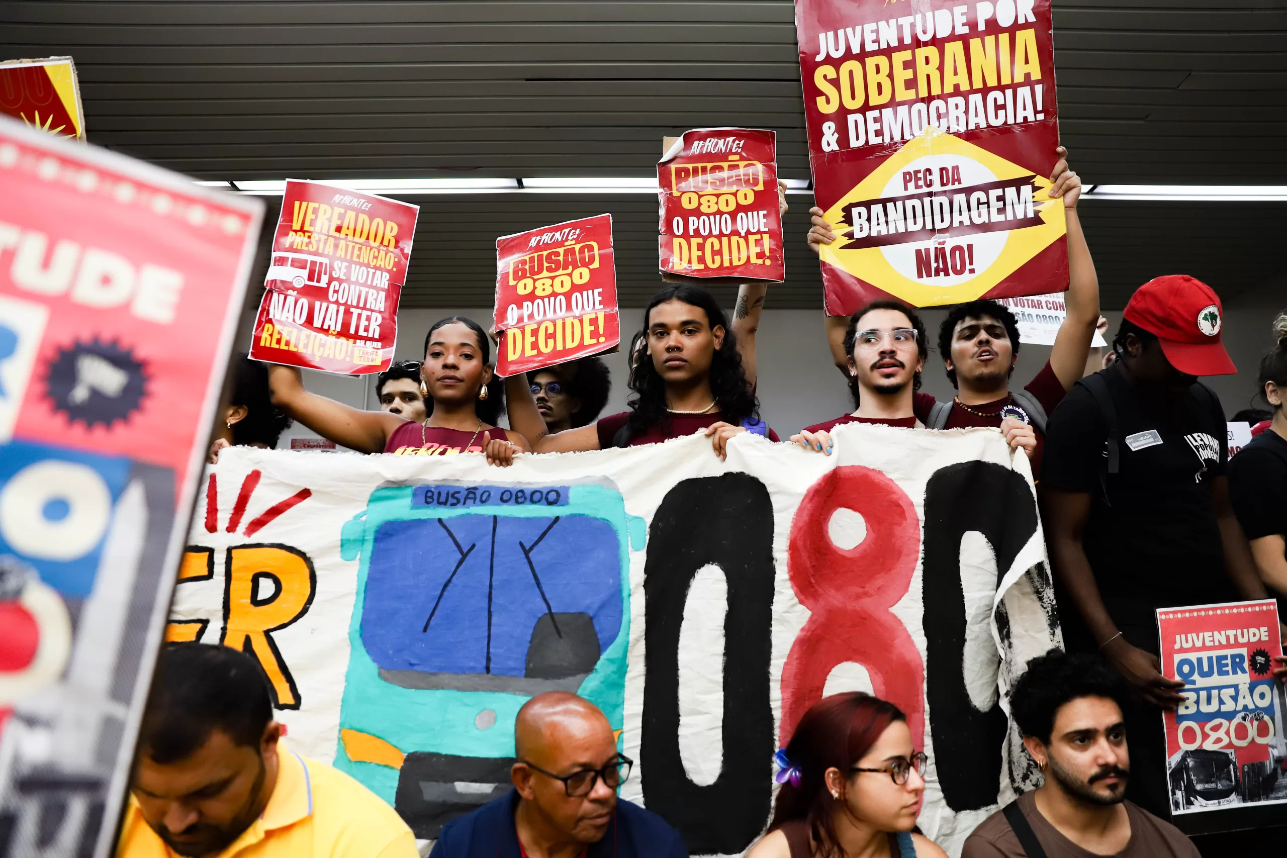 Grupo de manifestantes jovens seguram cartazes vermelhos e uma faixa com o desenho de um ônibus e o número “0800” (que significa, gratuidade), em protesto à favor do transporte gratuito e pela participação popular nas decisões públicas.