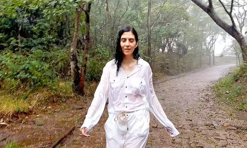 Camila, uma mulher branca e de cabelos pretos, no centro da imagem, vestida com uma roupa branca, tomando chuva em uma estrada de pedras e na chuva.