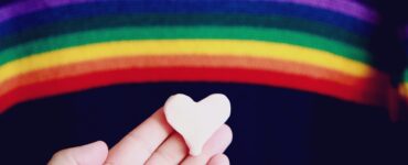 Bandeira LGBTQIA+ ao fundo, com uma mão segurando um coração