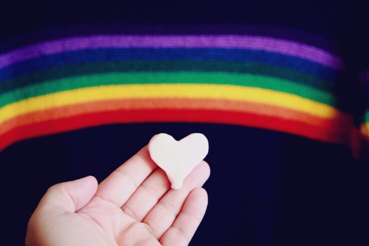 Bandeira LGBTQIA+ ao fundo, com uma mão segurando um coração