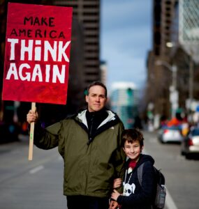 Homem com criança em manifestação segura placa vermelha com o escrito "Make America Think Again"