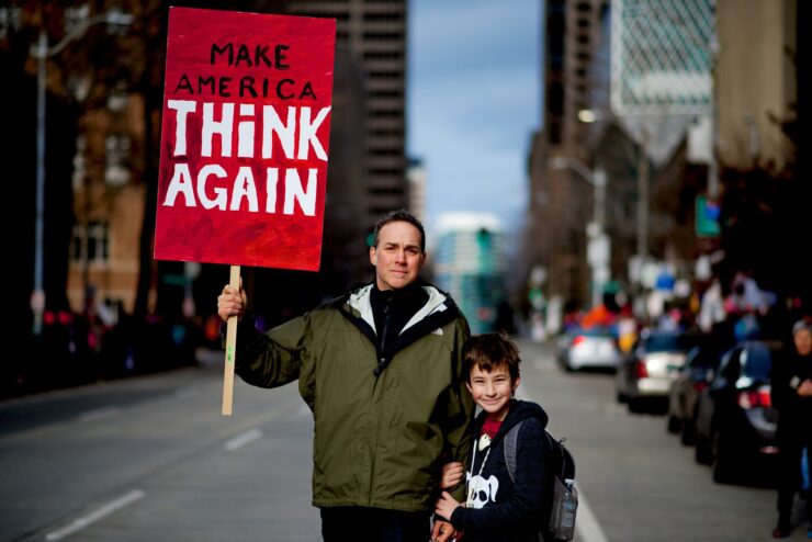 Homem com criança em manifestação segura placa vermelha com o escrito "Make America Think Again"