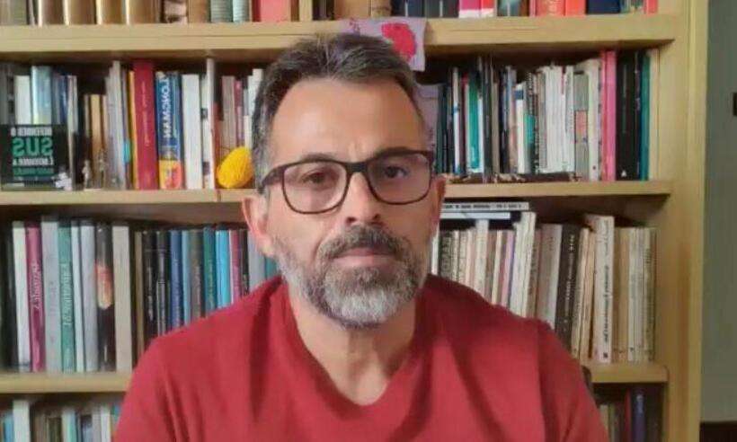 Professor Unaí Tupinambás é um homem branco de meia idade. Ele usa óculos, tem bigode, barba e cavanhaque e veste blusa vermelha. Ao fundo, uma estante de livros.