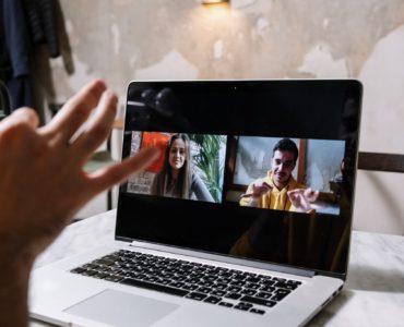 Uma pessoa branca utilizando Libras para se comunicar em uma videochamada. Há um notebook em uma mesa e ele mostra as duas pessoas que estão na videochamada: uma garota loira e um garoto com blusa amarela (esse garoto é o que está usando Libras)