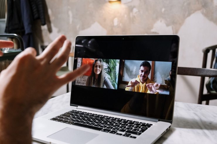 Uma pessoa branca utilizando Libras para se comunicar em uma videochamada. Há um notebook em uma mesa e ele mostra as duas pessoas que estão na videochamada: uma garota loira e um garoto com blusa amarela (esse garoto é o que está usando Libras)