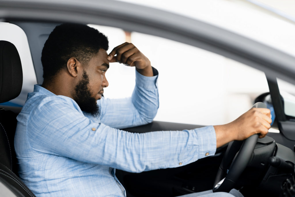 Motorista sentado no carro com uma mão na testa e expressão indicando cansaço.