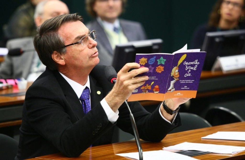 #paratodosverem Imagem de Bolsonaro segurando o livro “Aparelho sexual e cia” na Câmara dos Deputados. Jair aparece sentado na bancada e discursando a respeito do objeto em suas mãos