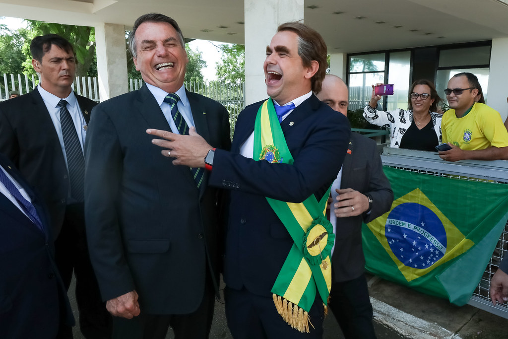 #paratodosverem Imagem de Bolsonaro abraçando um humorista vestido de terno e com a faixa presidencial. Os dois gargalham no momento da foto. Ao fundo, um segurança, apoiadores e uma bandeira do Brasil.