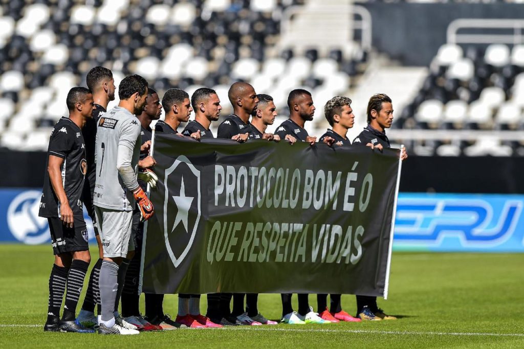 Time do Botafogo segura faixa nas cores do clube, no gramado antes do jogo, com o escudo do time e os dizeres “Protocolo bom é o que respeita vidas”.