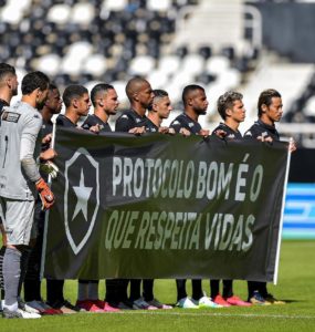 Time do Botafogo segura faixa nas cores do clube, no gramado antes do jogo, com o escudo do time e os dizeres “Protocolo bom é o que respeita vidas”.