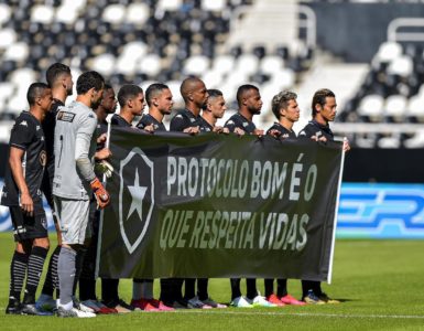 Time do Botafogo segura faixa nas cores do clube, no gramado antes do jogo, com o escudo do time e os dizeres “Protocolo bom é o que respeita vidas”.