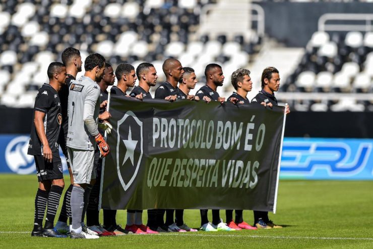 Time do Botafogo segura faixa nas cores do clube, no gramado antes do jogo, com o escudo do time e os dizeres “Protocolo bom é o que respeita vidas”.