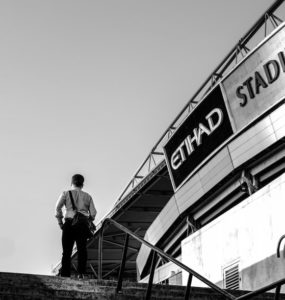 Foto em preto e branco com estádio da Etihad à direita. À esquerda, um homem de costas sobe escadas. Créditos: Phu Cuong Pham / Unsplash