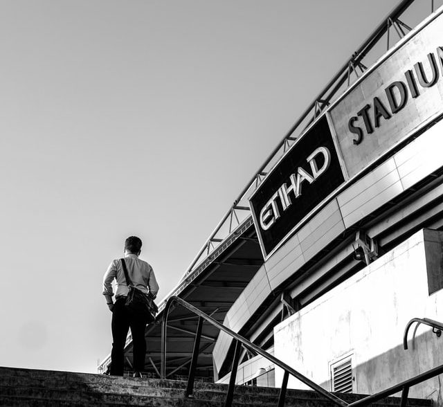 Foto em preto e branco com estádio da Etihad à direita. À esquerda, um homem de costas sobe escadas. Créditos: Phu Cuong Pham / Unsplash