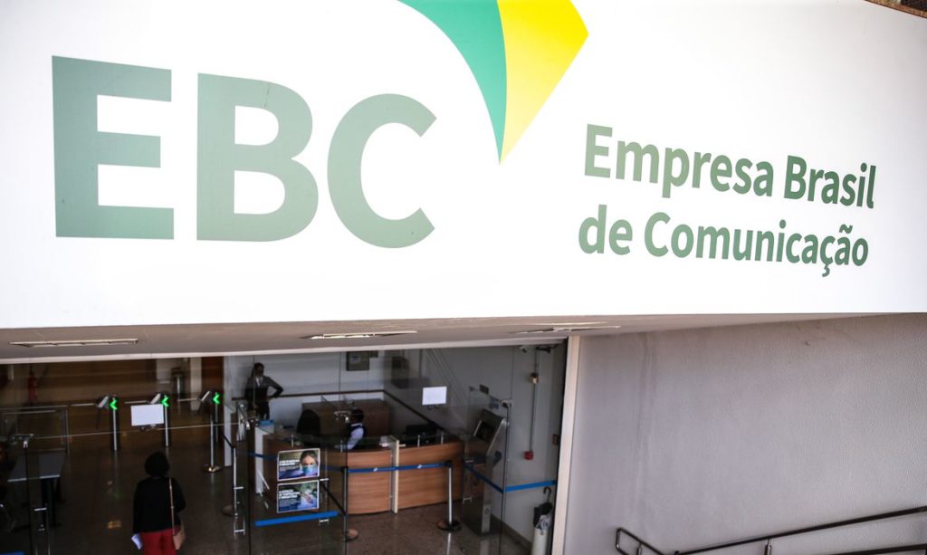 Fachada da Empresa Brasil de Comunicação. Prédio branco escrito em destaque no canto esquerdo EBC na cor verde, ao centro um desenho verde e amarelo e no canto direito está escrito Empresa Brasil de Comunicação na cor verde. Na imagem há também o hall de entrada do prédio, onde contém catracas, bancada de madeira e pessoas circulando no local.