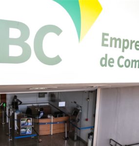 Fachada da Empresa Brasil de Comunicação. Prédio branco escrito em destaque no canto esquerdo EBC na cor verde, ao centro um desenho verde e amarelo e no canto direito está escrito Empresa Brasil de Comunicação na cor verde. Na imagem há também o hall de entrada do prédio, onde contém catracas, bancada de madeira e pessoas circulando no local.