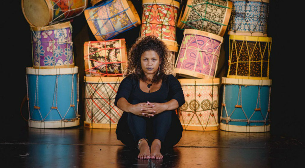 Em um palco com tambores coloridos empilhados ao fundo, Júlia Tizumba, mulher negra, se senta vestida de preto. Ela apoia as mãos nos joelhos e tem um semblante sério e sereno.