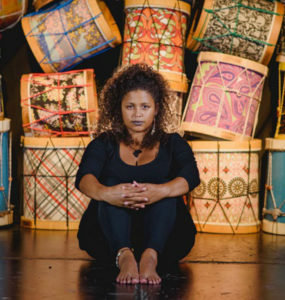 Em um palco com tambores coloridos empilhados ao fundo, Júlia Tizumba, mulher negra, se senta vestida de preto. Ela apoia as mãos nos joelhos e tem um semblante sério e sereno.