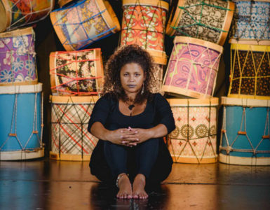 Em um palco com tambores coloridos empilhados ao fundo, Júlia Tizumba, mulher negra, se senta vestida de preto. Ela apoia as mãos nos joelhos e tem um semblante sério e sereno.