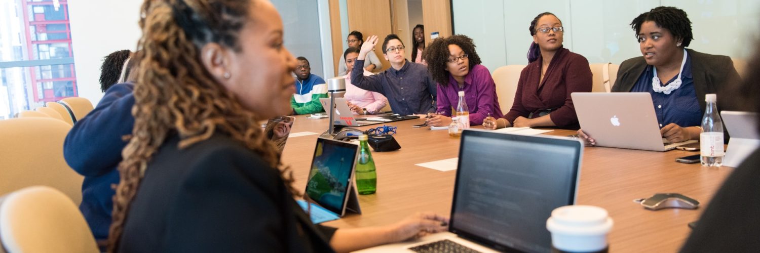 Um grupo diverso de mulheres negras e pretas se reúne em torno de uma mesa de madeira em ambiente corporativo