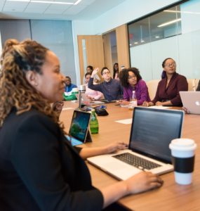 Um grupo diverso de mulheres negras e pretas se reúne em torno de uma mesa de madeira em ambiente corporativo