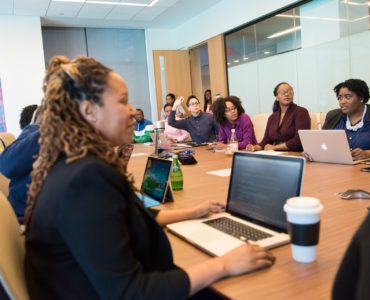 Um grupo diverso de mulheres negras e pretas se reúne em torno de uma mesa de madeira em ambiente corporativo