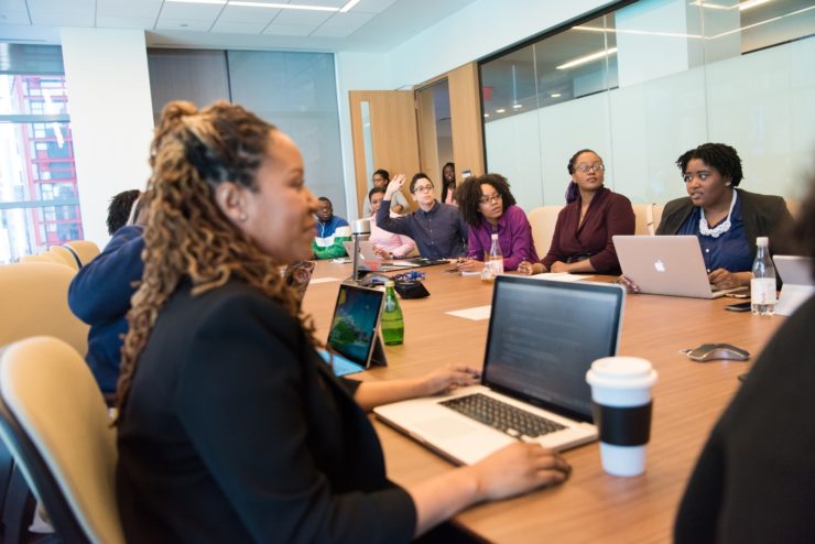 Um grupo diverso de mulheres negras e pretas se reúne em torno de uma mesa de madeira em ambiente corporativo