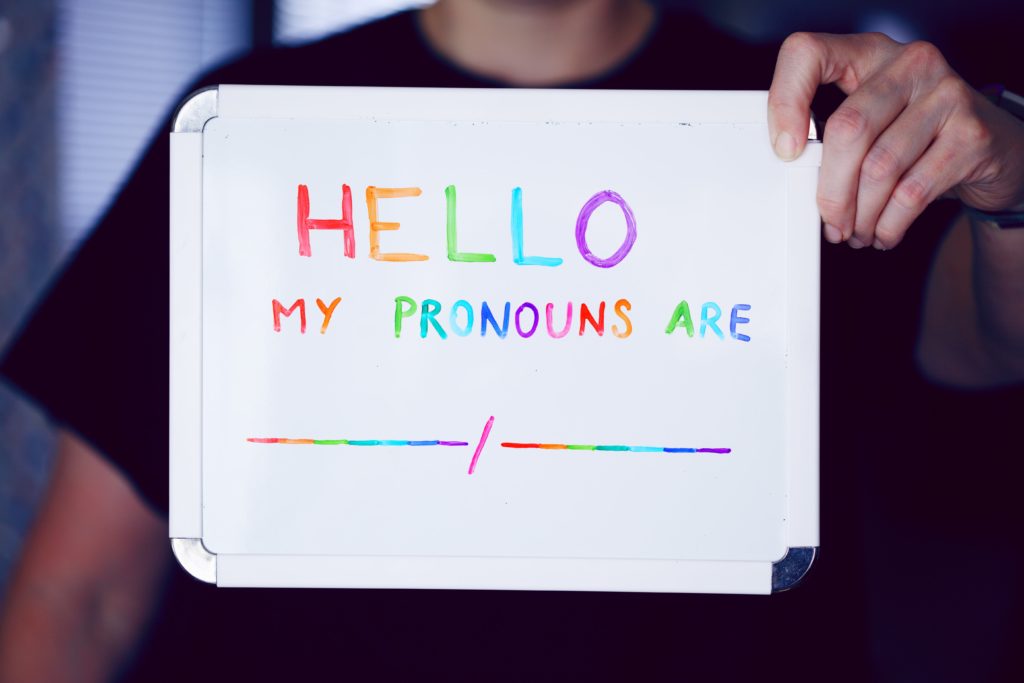 Uma pessoa branca veste uma camisa preta e segura um pequeno quadro branco em que se lê: "Hello, my pronouns are" / "Olá, meus pronomes são" (em tradução livre). A imagem da pessoa está desfocada e a mensagem está escrita com as cores do arco-íris.