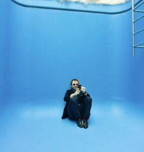 homem branco usando roupa preta sentado no no chão de obra que representa um fundo de uma piscina, na cor azul