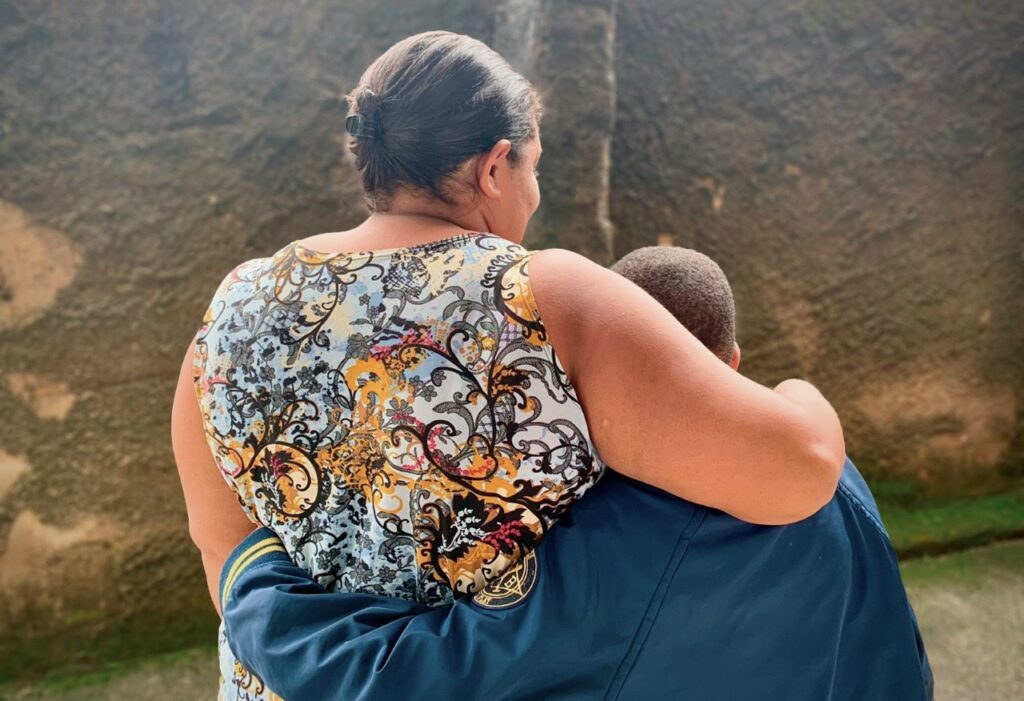 Mulher de costas, à esquerda, com blusa estampata nos tons branco, preto, amarelo e vermelho abraça filho, à direita, também de costas, com jaqueta azul escuta. Ambos estão virados para um muro de cimento marcado pelo tempo e musgo. A mulher tem o rosto levemente virado à direita, o que permite ver que ela está sorrindo.