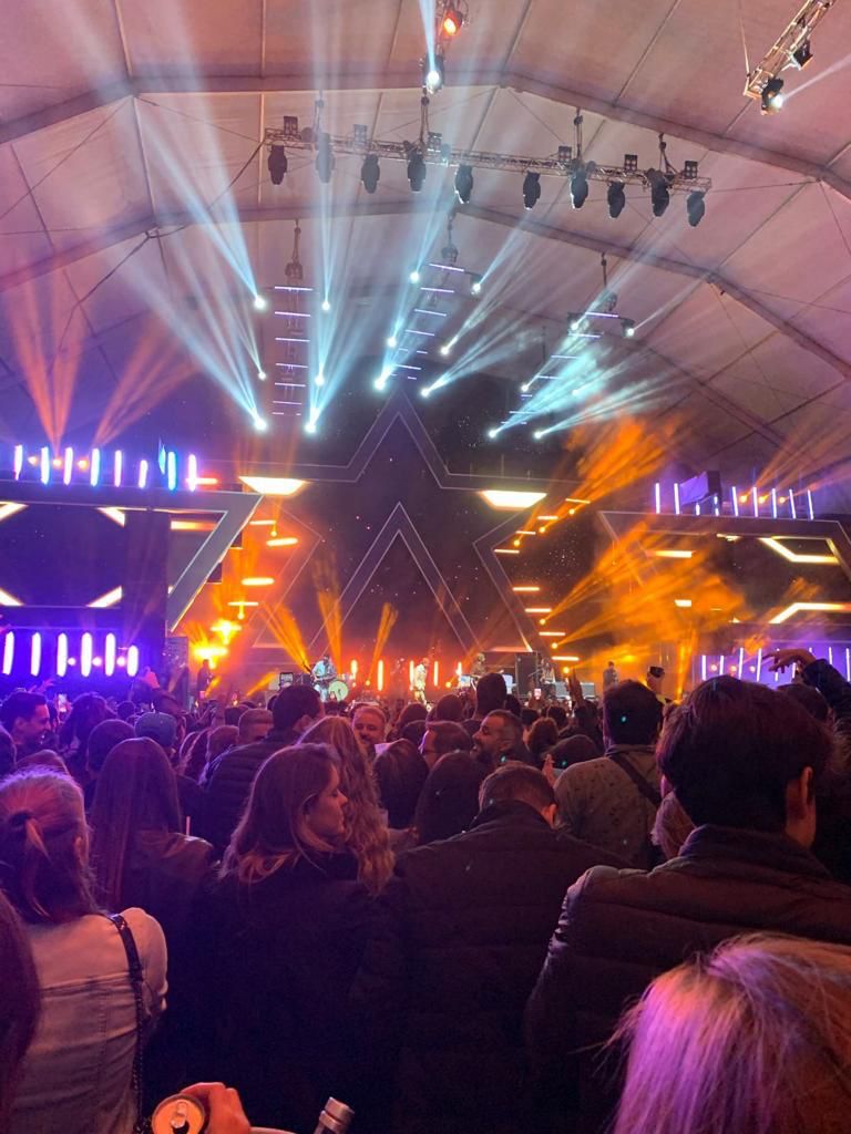 Visão geral do público frente ao palco com a estrutura de estrela, todo iluminado pelas cores azul, branco e laranja. No centro, a banda em cima do palco.