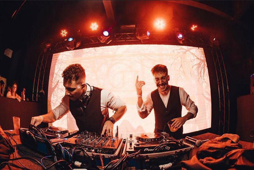 Os artistas Pedro Braichi e Narder DJ estão em foco na cena, atrás da mesa de DJ, eles sorriem, enquanto um toca o aparelho e o outro segura um copo e levanta a mão. A estrutura do palco em que os artistas estão é iluminada e na lateral do ambiente está uma parte do público.