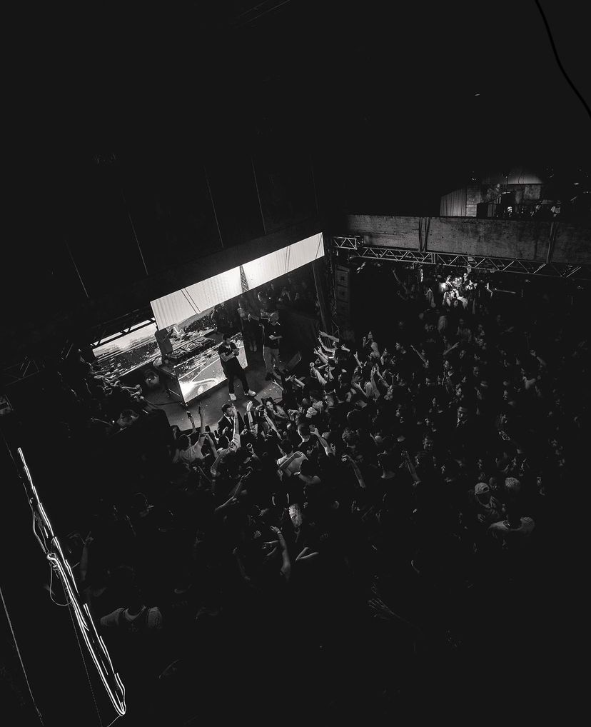 Foto em preto e branco, visão geral pelo ângulo de cima que mostra o público de frente para o palco. No palco os artistas interagem com as pessoa, enquanto um canta o outro tira uma selfie com alguém da plateia.