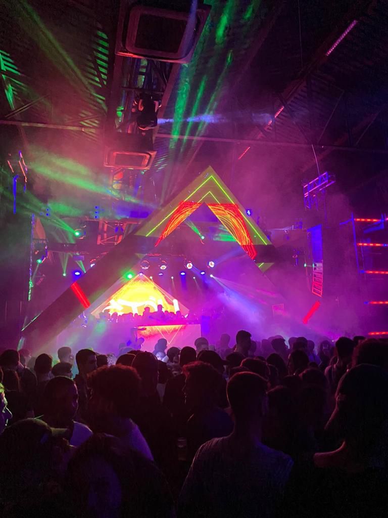 No palco o DJ, seus dançarinos e algumas pessoas da plateia se juntam atrás da cabine. As luzes do palco nas cores verde, azul, laranja e roxo iluminam o ambiente a frente em que o público assiste o show.