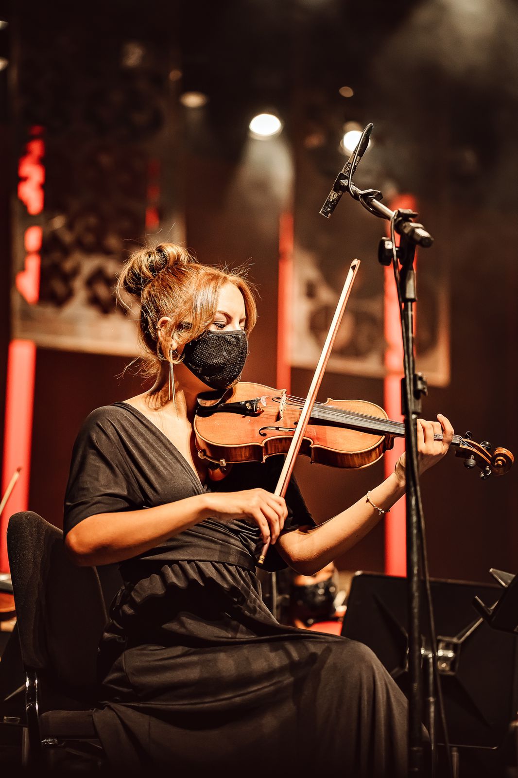 Apenas a violinista, o violino, o microfone e seu pedestal em foco na cena. De roupa e máscara preta, a violinista sentada toca seu violino marrom. À sua frente o microfone e pedestal, já no fundo, em desfoque, parte do ambiente do palco.