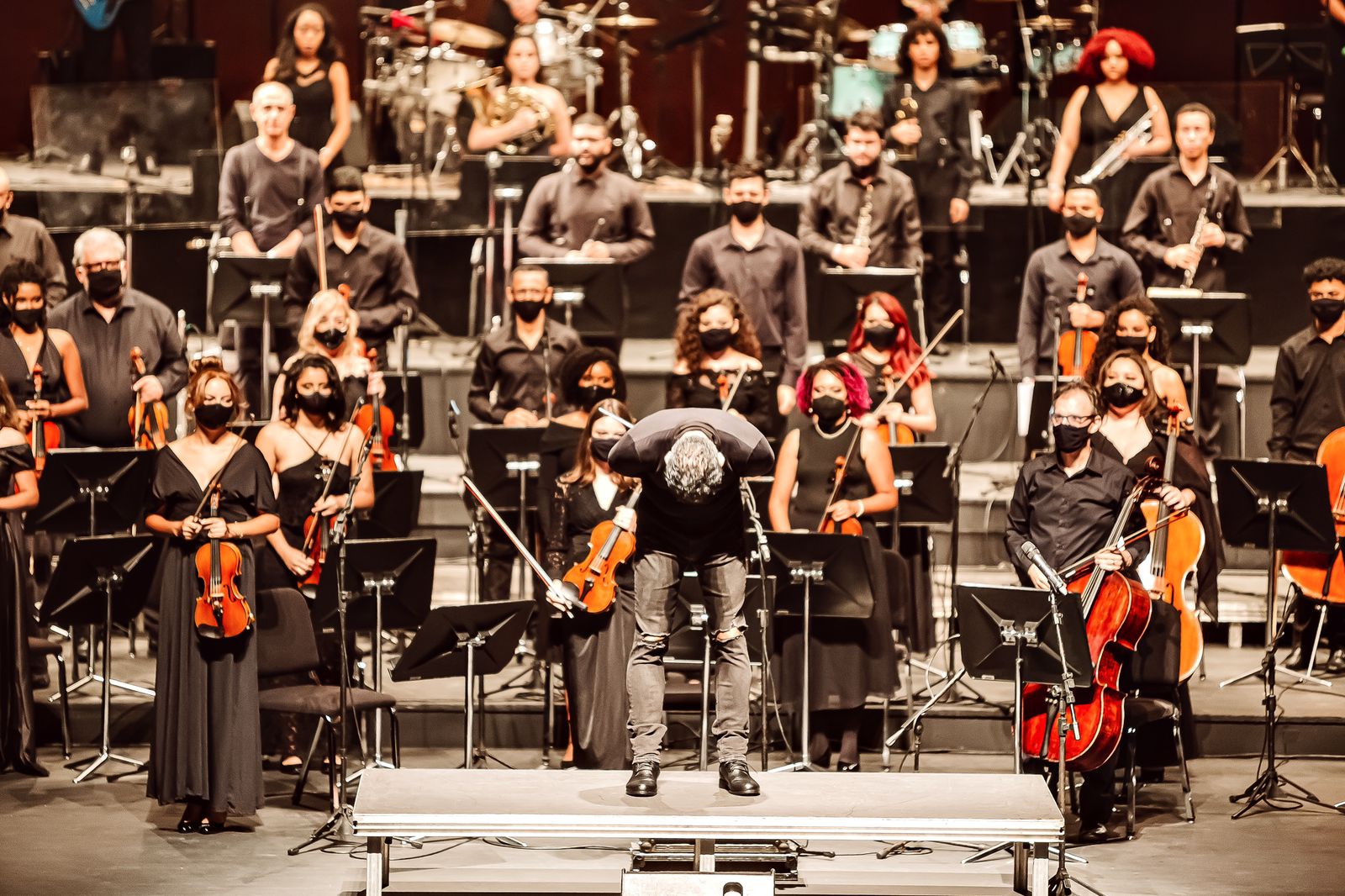 Toda orquestra de máscara, em cima do palco, segura seus instrumentos. O maestro se curva em agradecimento ao público que não aparece na imagem.