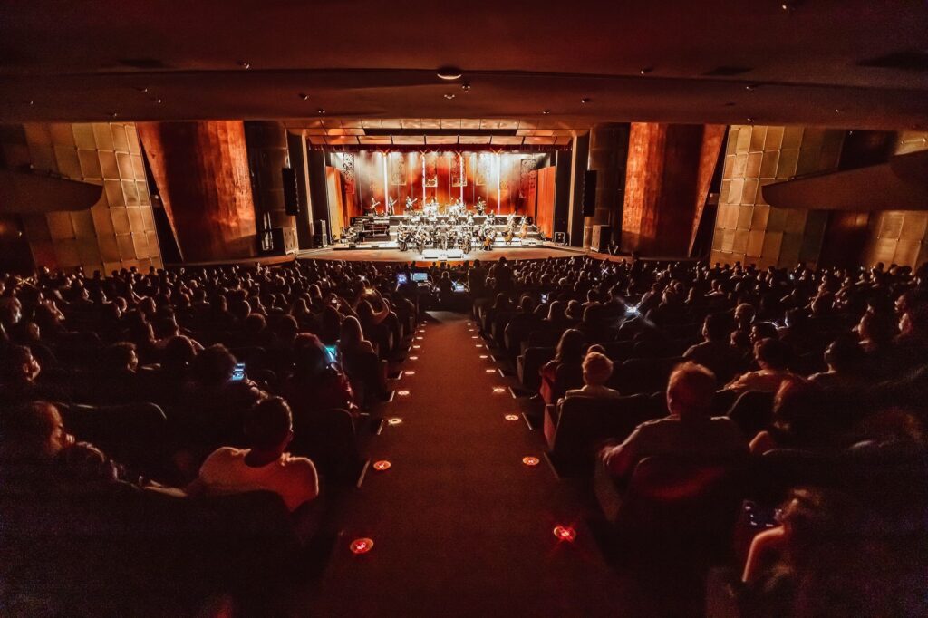 Com foco no público, está visão de trás do teatro que no fundo mostra o palco que iluminado, abriga a orquestra.