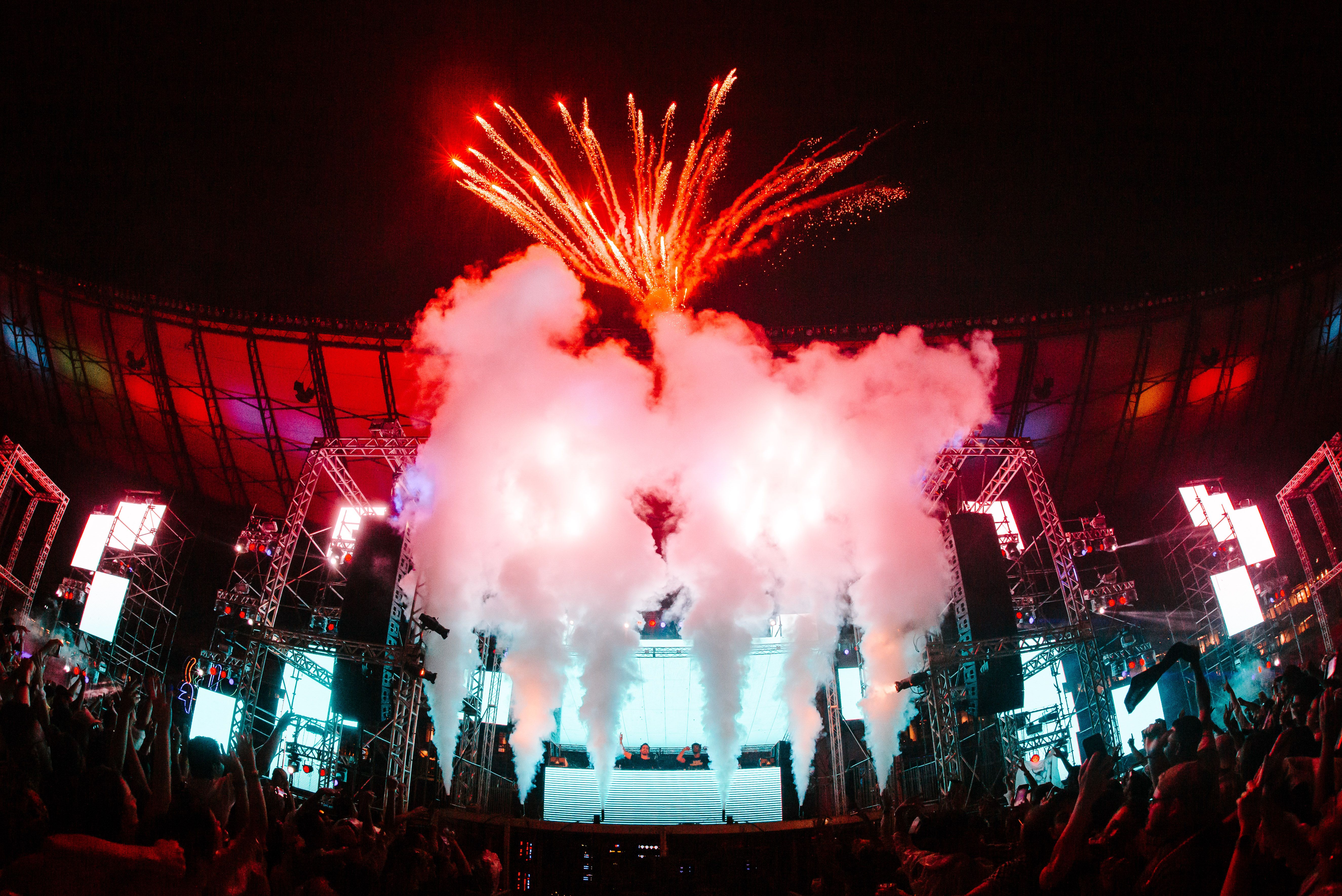 Imagem mostra palco de um show de eletrônica no estádio Mineirão. Há pessoas com as mãos levantadas para cima e fogos de artifício no céu