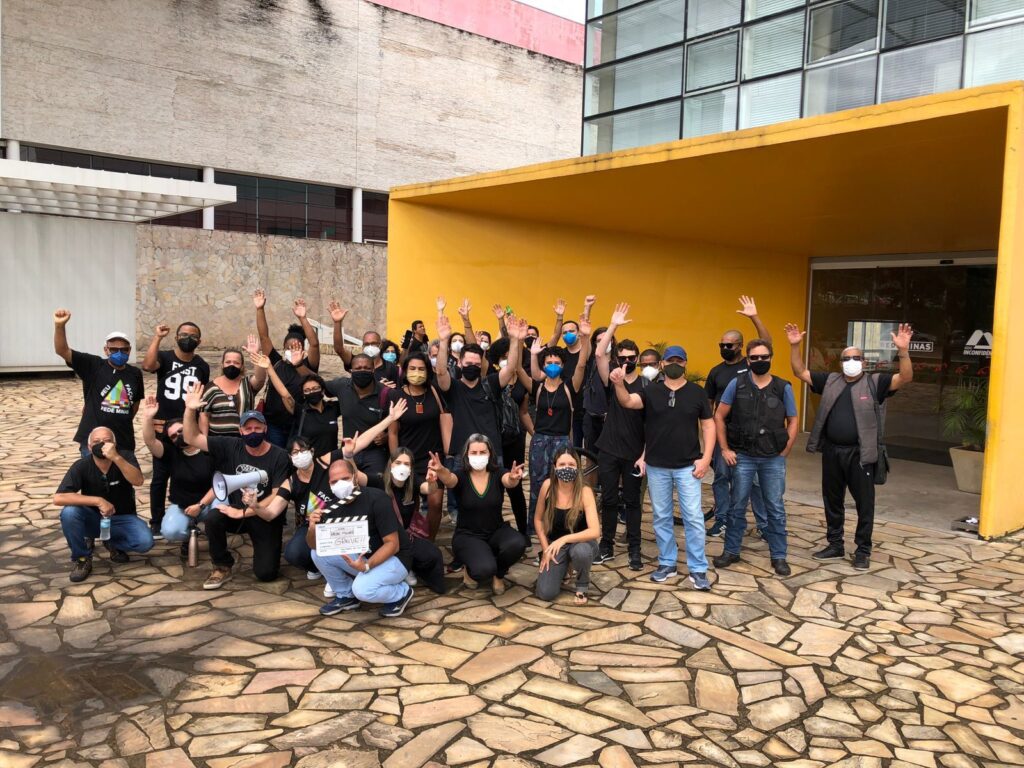 Grupo de pessoas vestidas de preto, em frente a um prédio de entrada amarela e janelas de vidro