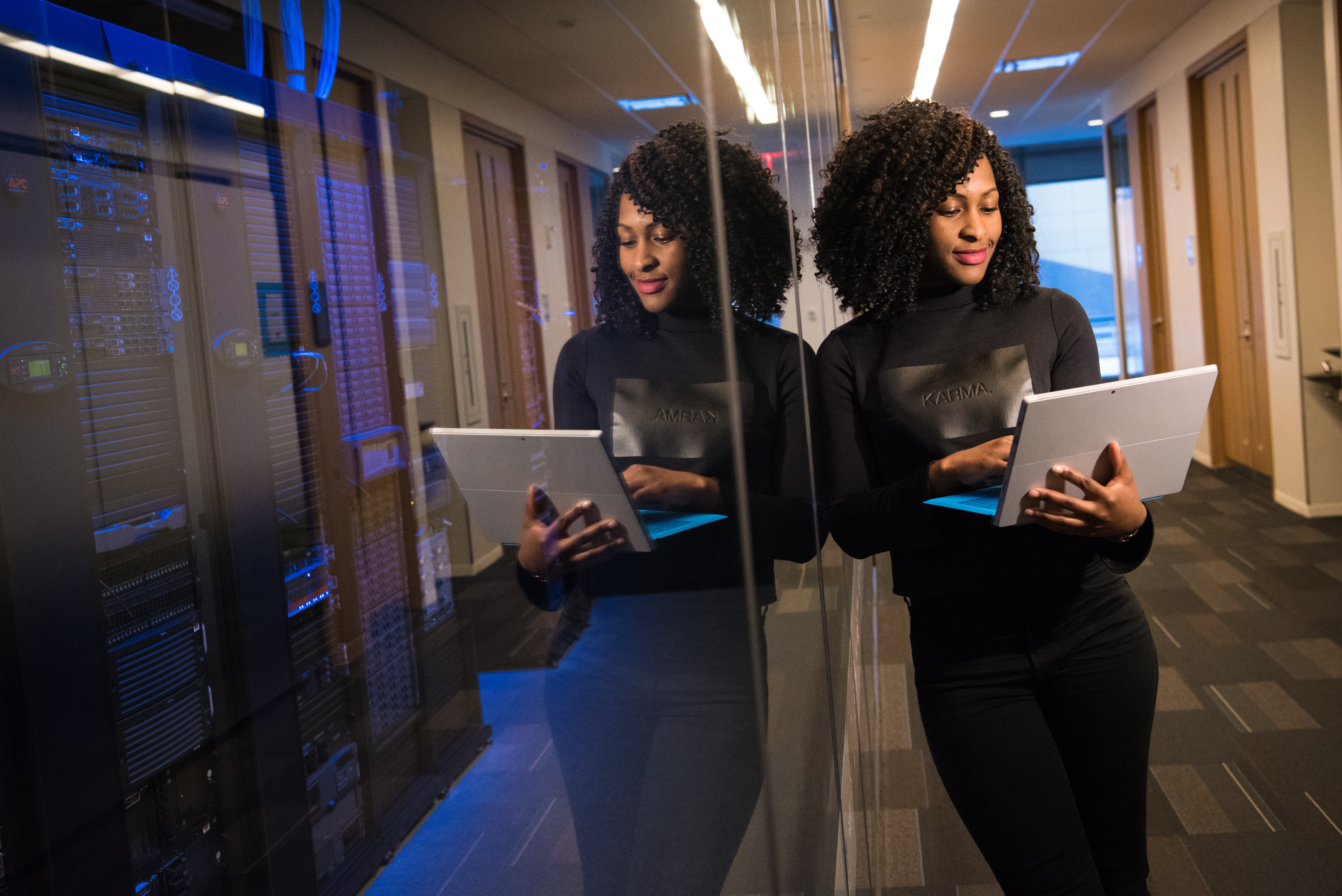 mulher negra inclinada sobre vidro segurando um computador no corredor de uma empresa