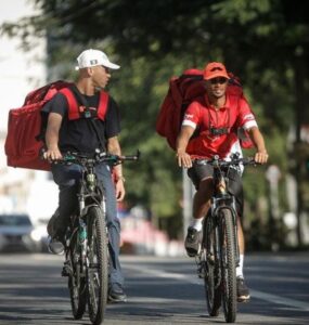 Dois entregadores de aplicativo, andando de bicicleta na rua e conversando.