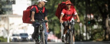 Dois entregadores de aplicativo, andando de bicicleta na rua e conversando.