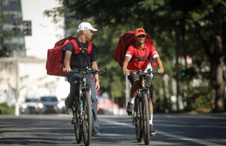 Dois entregadores de aplicativo, andando de bicicleta na rua e conversando.