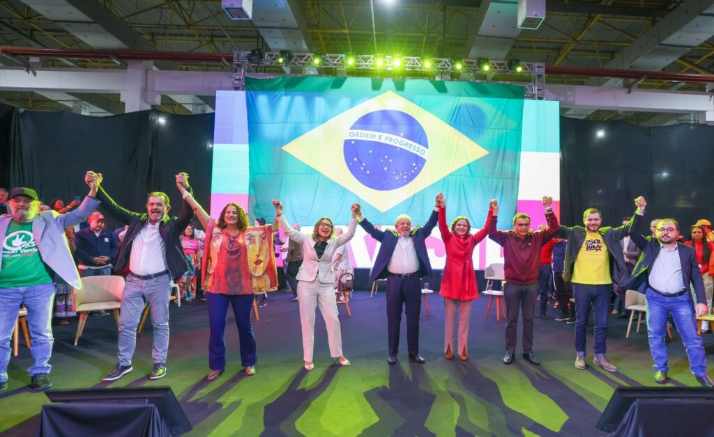 Foto do evento de pré-lançamento da candidatura de Lula. Foto mostra o candidato de mãos dadas com aliados, entre eles sua esposa janja, em frente a um telão que exibe a bandeira do Brasil 