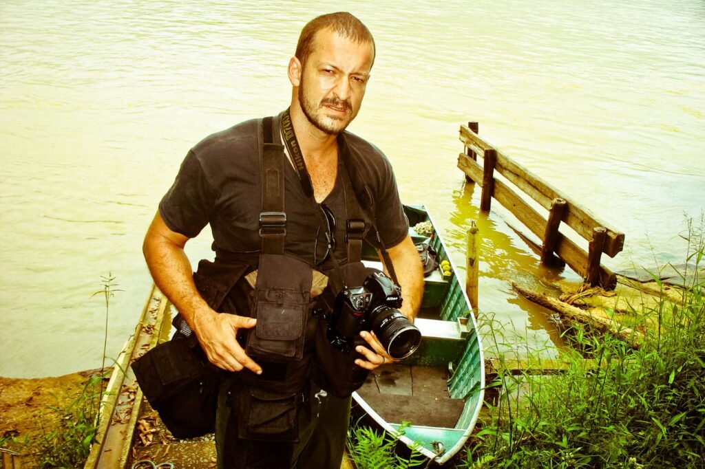 Leandro Couri parado olhando para a câmera frente a um lago segurando uma câmera fotográfica.