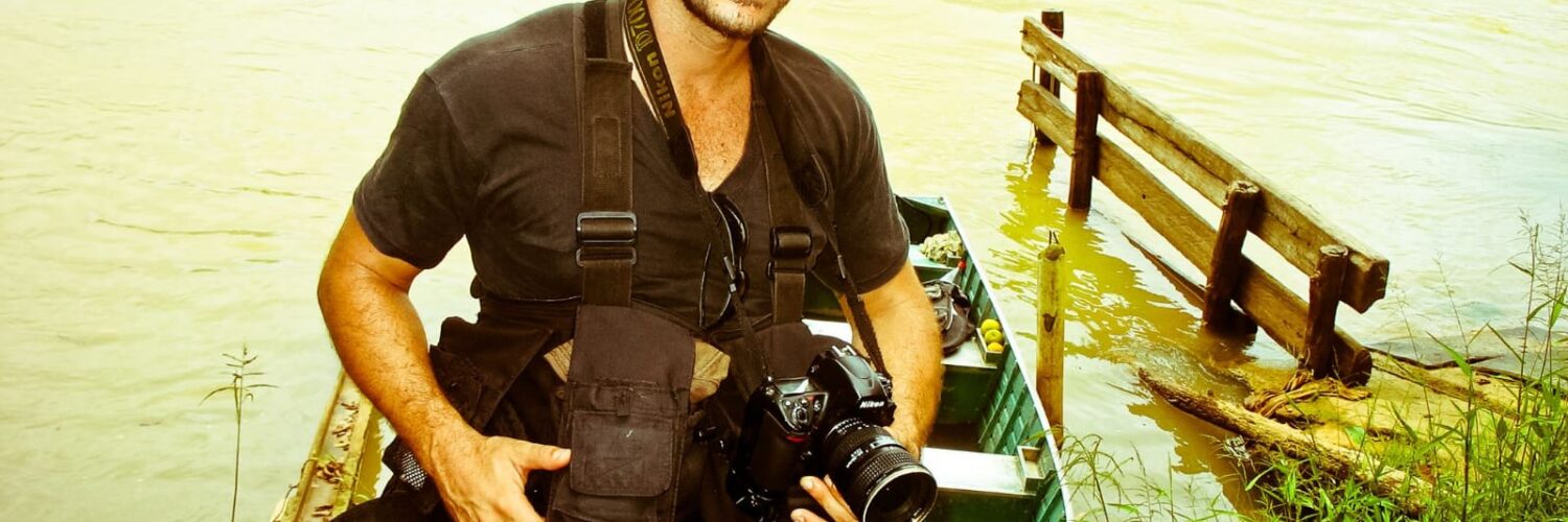 Leandro Couri parado olhando para a câmera frente a um lago segurando uma câmera fotográfica.