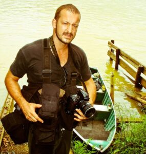 Leandro Couri parado olhando para a câmera frente a um lago segurando uma câmera fotográfica.