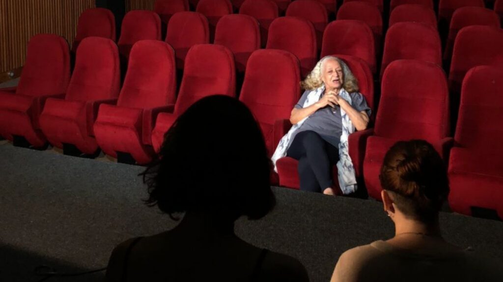 A imagem está dividida em dois planos: um com as costas das cineastas em formação, em primeiro plano, e o segundo com uma arquibancada de cinema na cor vermelha com uma atriz senhora de cabelos brancos conversando, que representa a Dona.