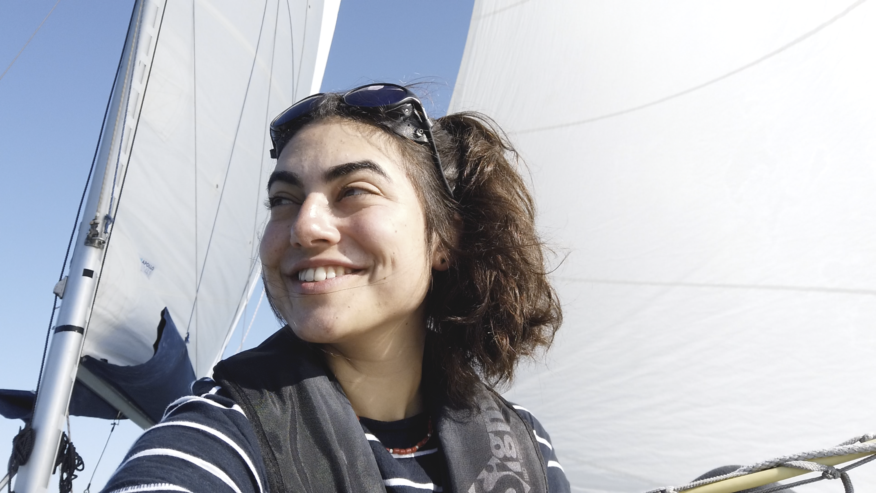 Tamara Klink (branca de cabelos castanhos) olha para o lado esquerdo enquanto senta em seu barco. Ao fundo, aparecem as velas.