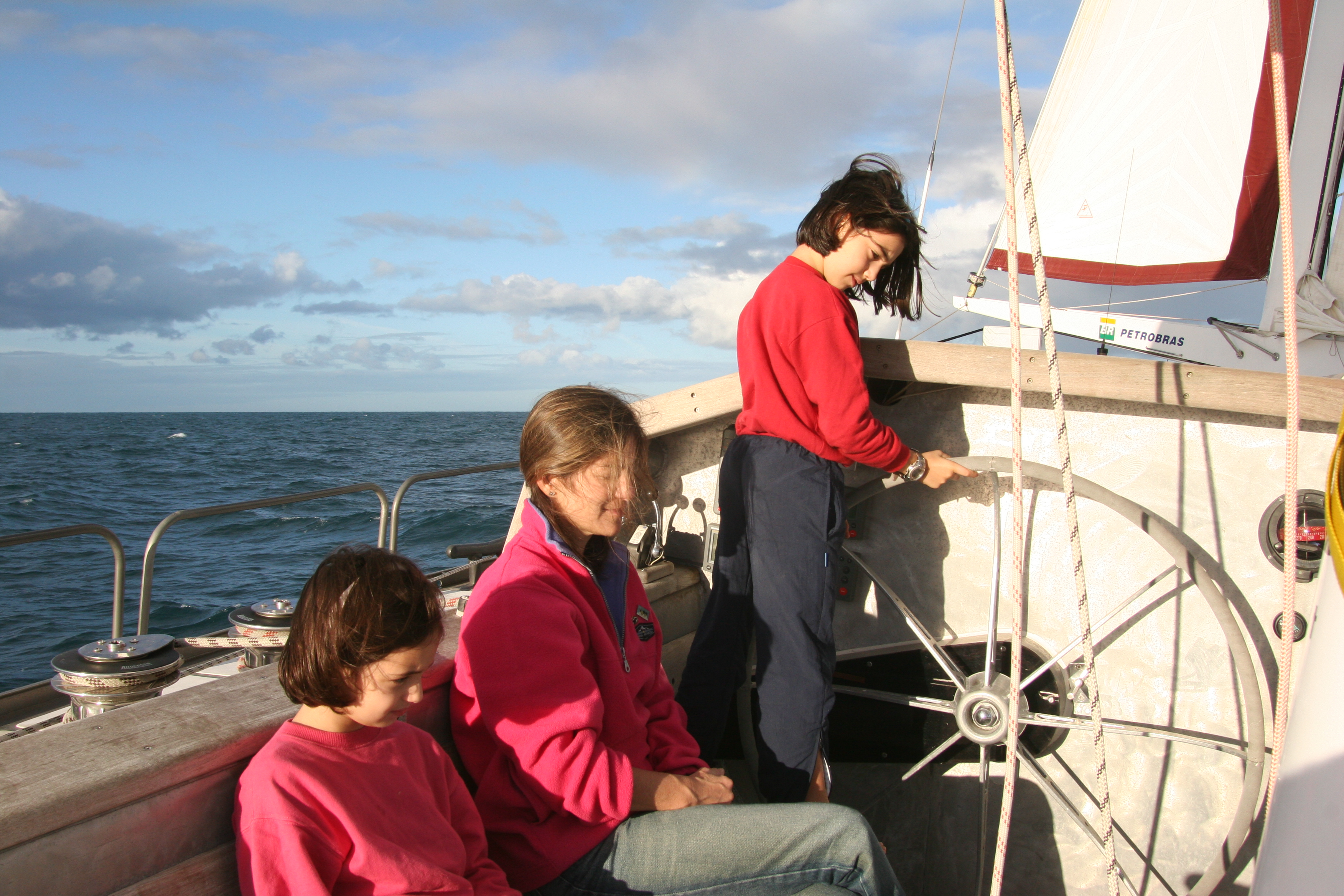 Tamara, sua mãe e sua irmã vestem jaquetas vermelhas em um barco. Tamara está em pé e mexe no leme. Sua mãe e sua irmã estão sentadas. Faz sol. O barco é branco. 